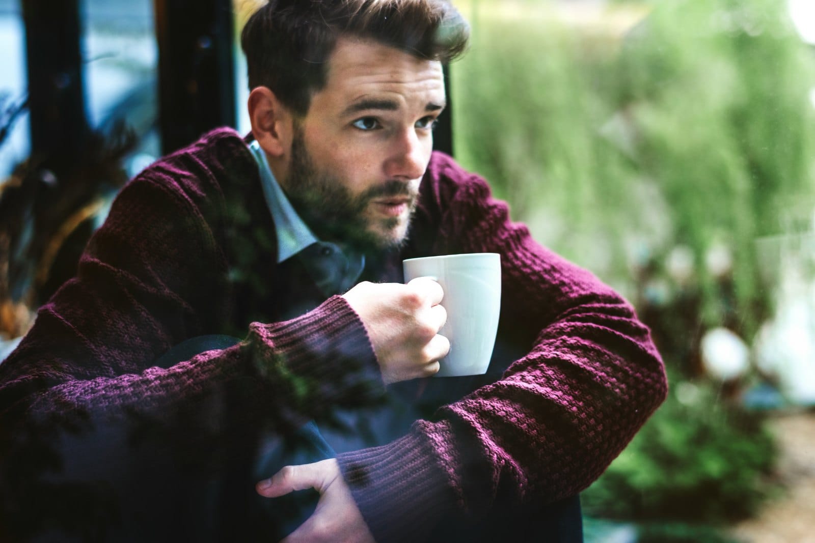 A bearded man in a dark red knit sweater holding a mug, seated by a window
