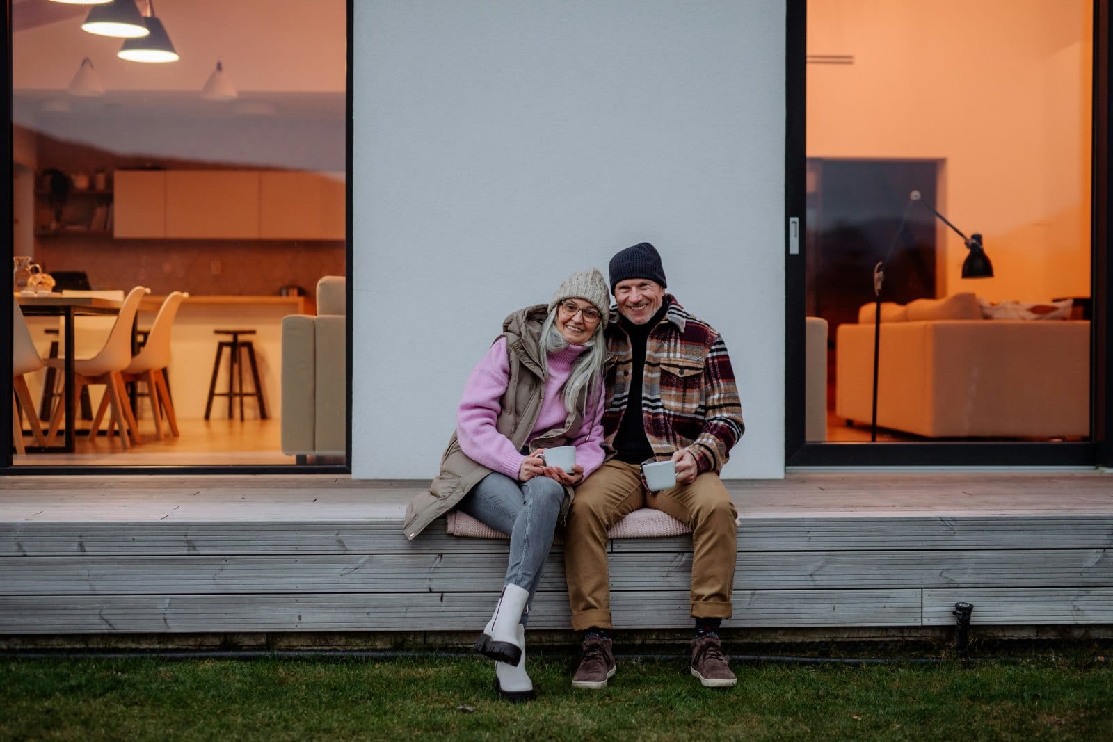 A couple in their sixties sitting outside a modern house holding a mug each, with lit rooms visible on either side
