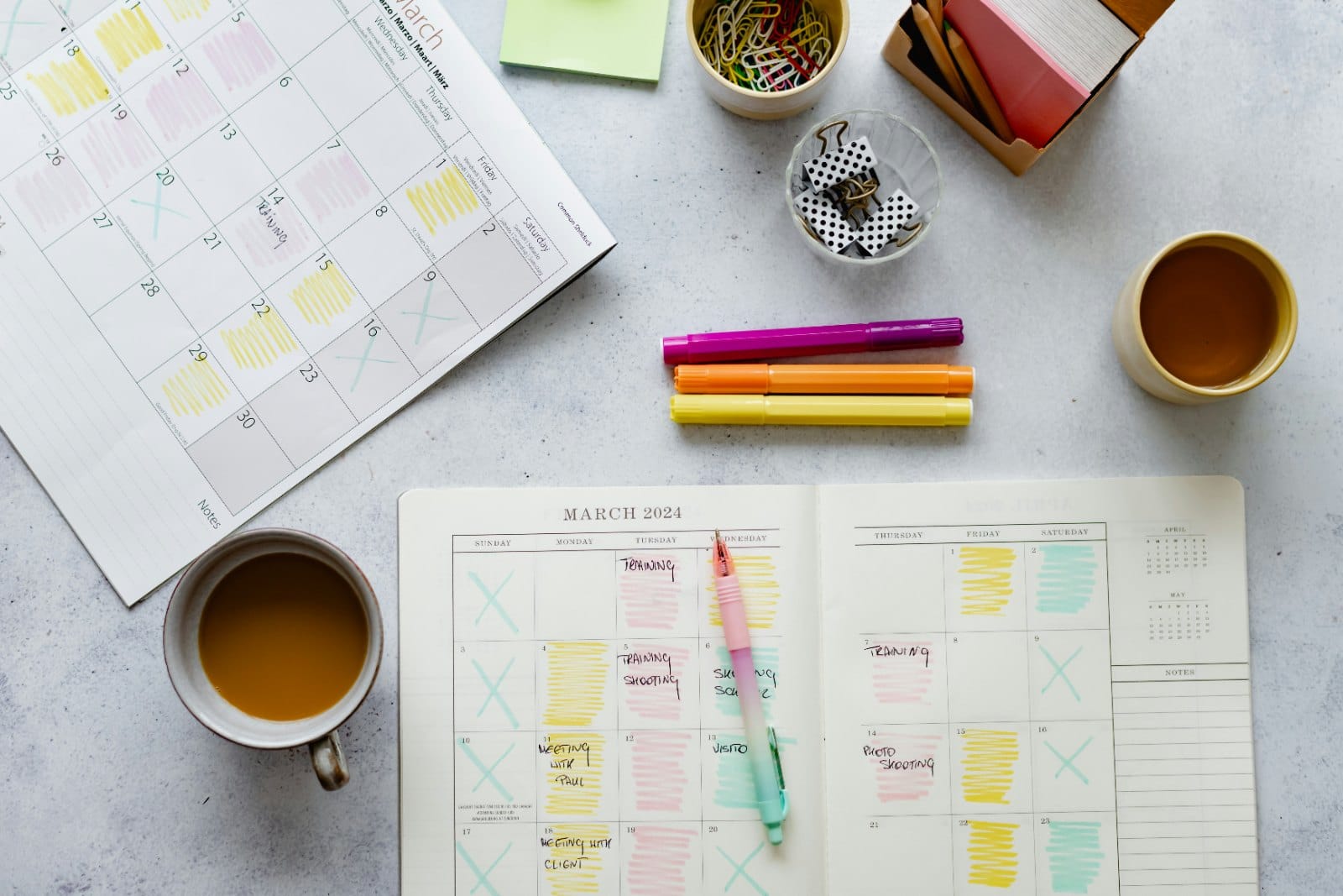 A monthly planner marked with highlighter, coloured pens and a cup of coffee on a desk
