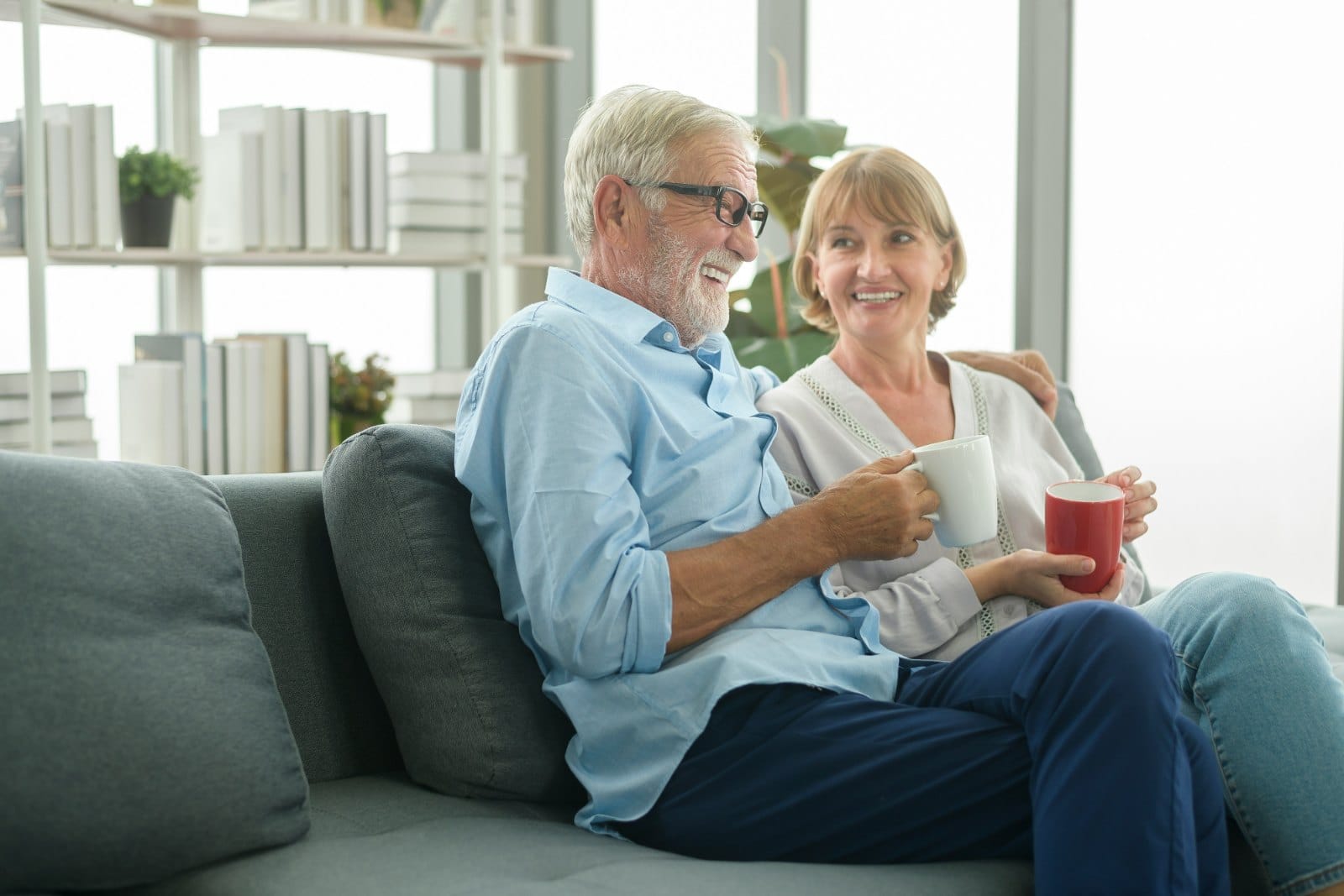 A couple in their sixties sitting together on a sofa with mugs of coffee, laughing