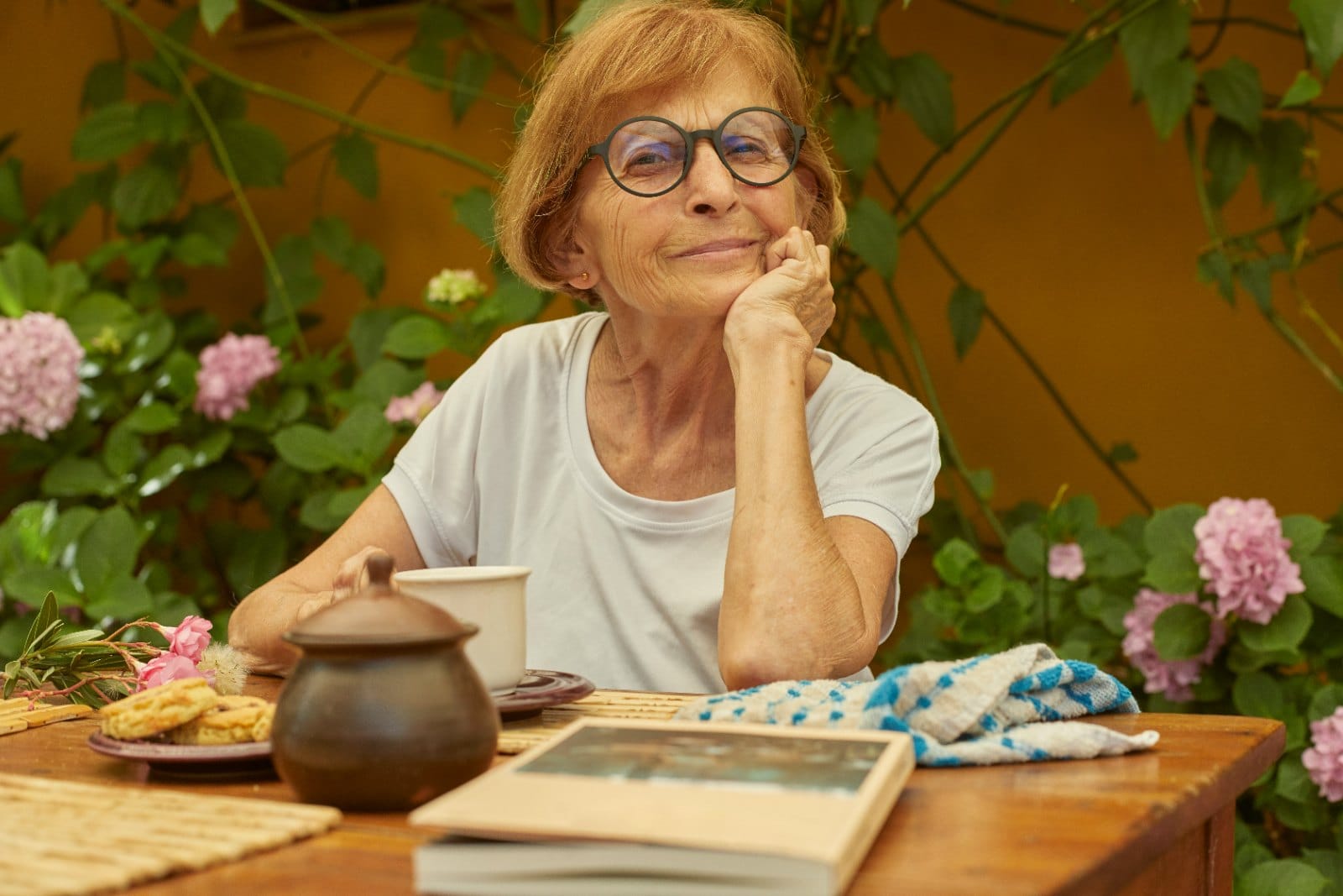 A woman sitting at an outdoor table with a teapot, a book and flowering plants behind her
