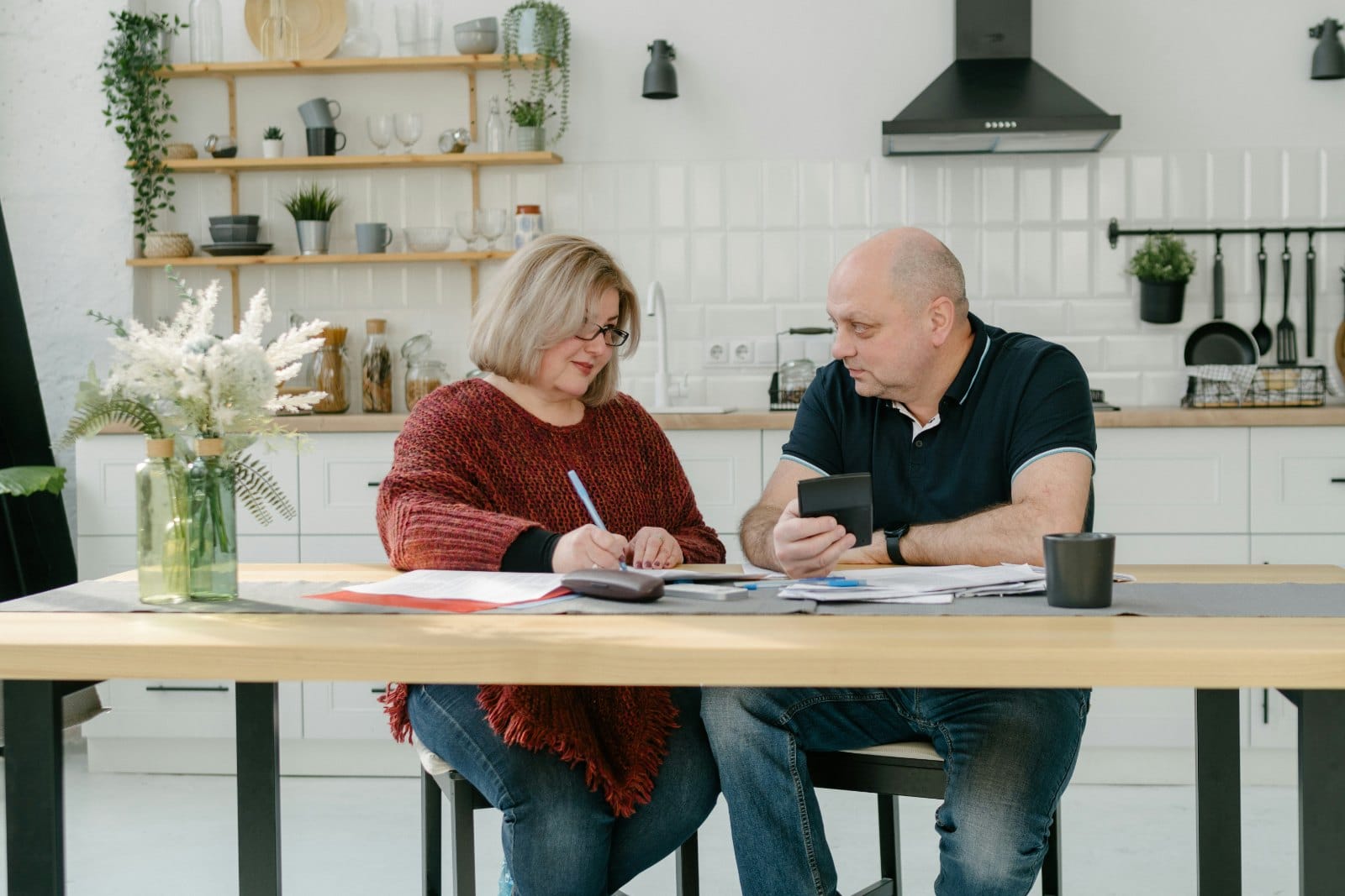 A couple in their fifties sitting at a kitchen table with paperwork and a calculator between them
