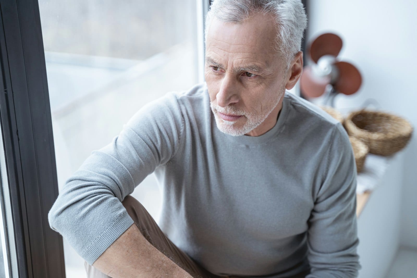 A man in his sixties sitting by a window, looking thoughtful
