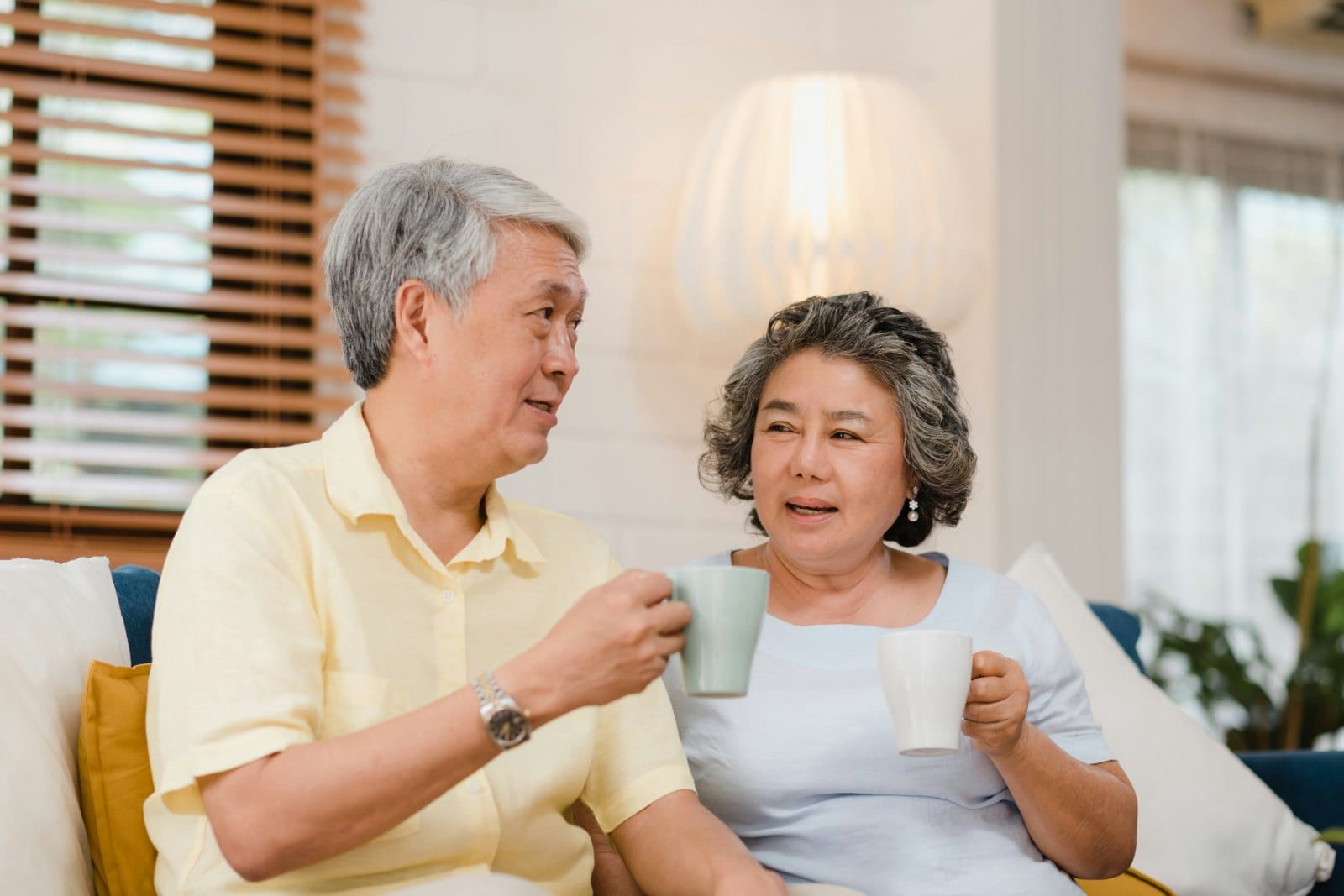 An older couple sitting together on a sofa with mugs of coffee, talking